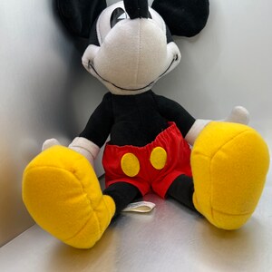 May include: A plush Mickey Mouse toy with black ears, a black body, and a white face. The toy wears red shorts with yellow buttons and large yellow gloves. The toy is sitting down.