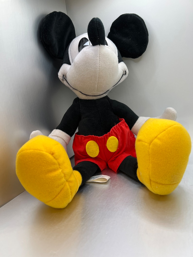 May include: A plush Mickey Mouse toy with black ears, a black body, and a white face. The toy wears red shorts with yellow buttons and large yellow gloves. The toy is sitting down.