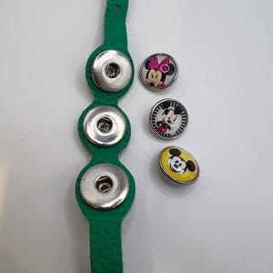 Puede incluir: Una correa de cuero verde con tres cierres de bot&oacute;n a presi&oacute;n plateados. Tres botones decorativos a presi&oacute;n presentan dise&ntilde;os de Mickey Mouse y Minnie Mouse en negro, blanco, amarillo y rosa. Los botones son redondos.