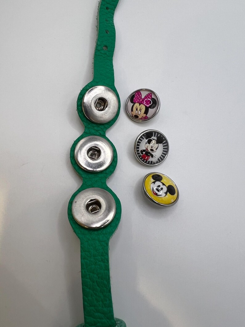 Puede incluir: Una correa de cuero verde con tres cierres de bot&oacute;n a presi&oacute;n plateados. Tres botones decorativos a presi&oacute;n presentan dise&ntilde;os de Mickey Mouse y Minnie Mouse en negro, blanco, amarillo y rosa. Los botones son redondos.