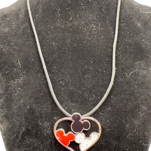 Puede incluir: Un collar con un colgante de coraz&oacute;n plateado con tres cabezas de Mickey Mouse en su interior. Las cabezas de Mickey Mouse son rojas, negras y blancas con diamantes de imitaci&oacute;n.