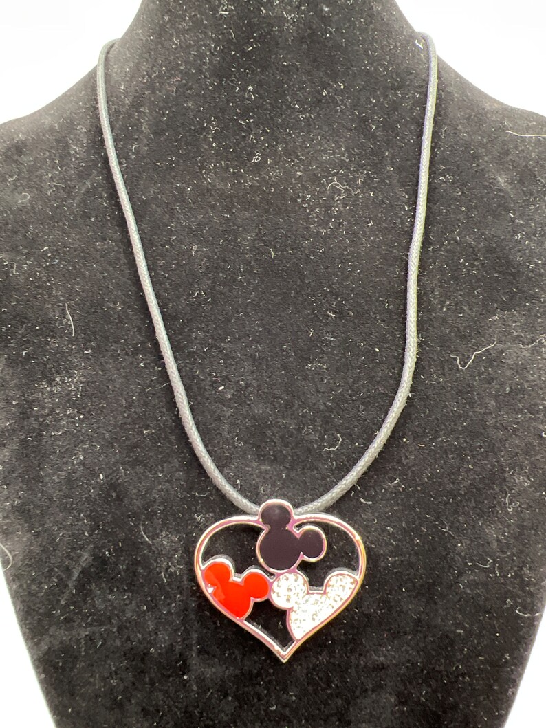Puede incluir: Un collar con un colgante de coraz&oacute;n plateado con tres cabezas de Mickey Mouse en su interior. Las cabezas de Mickey Mouse son rojas, negras y blancas con diamantes de imitaci&oacute;n.