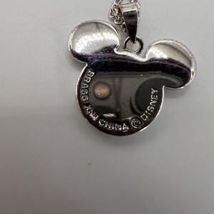 K&ouml;nnte beinhalten: Silberfarbene Halskette mit einem Mickey Mouse-Anh&auml;nger. Der Anh&auml;nger hat die Form von Mickys Kopf mit zwei gro&szlig;en Ohren. Auf der R&uuml;ckseite des Anh&auml;ngers steht "BRASS", "MADE IN CHINA" und "DISNEY".