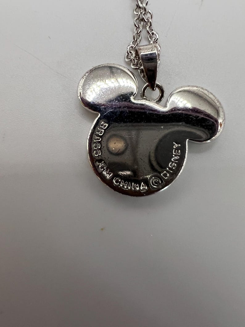 K&ouml;nnte beinhalten: Silberfarbene Halskette mit einem Mickey Mouse-Anh&auml;nger. Der Anh&auml;nger hat die Form von Mickys Kopf mit zwei gro&szlig;en Ohren. Auf der R&uuml;ckseite des Anh&auml;ngers steht "BRASS", "MADE IN CHINA" und "DISNEY".