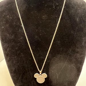 K&ouml;nnte beinhalten: Silberfarbene Halskette mit einem Anh&auml;nger in Form eines Mickey Mouse-Kopfes. Der Anh&auml;nger ist mit einem strukturierten, glitzernden Material gef&uuml;llt. Die Halskette wird vor einem schwarzen Hintergrund pr&auml;sentiert.