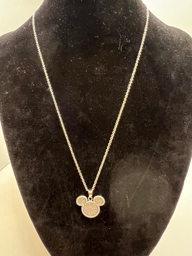 K&ouml;nnte beinhalten: Silberfarbene Halskette mit einem Anh&auml;nger in Form eines Mickey Mouse-Kopfes. Der Anh&auml;nger ist mit einem strukturierten, glitzernden Material gef&uuml;llt. Die Halskette wird vor einem schwarzen Hintergrund pr&auml;sentiert.