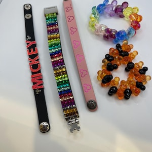 Puede incluir: Una colecci&oacute;n de pulseras de varios estilos. Una pulsera de cuero negro con letras rojas "MICKEY", una pulsera de diamantes de imitaci&oacute;n arco&iacute;ris, una pulsera de cuero rosa con siluetas de Mickey Mouse y pulseras de cuentas en arco&iacute;ris y naranja y negro.
