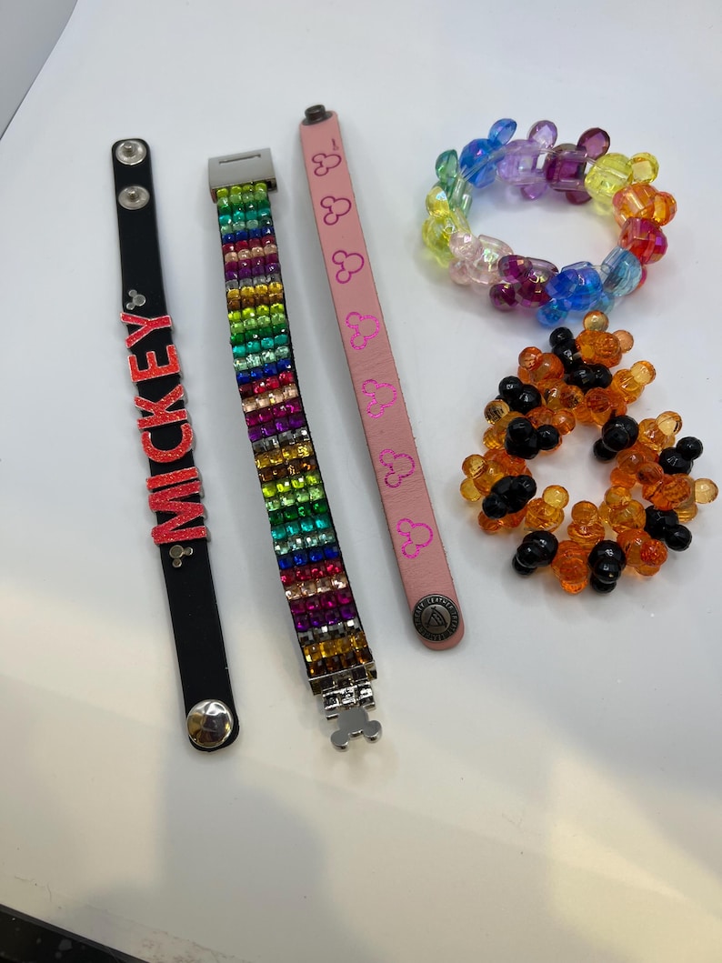 Puede incluir: Una colecci&oacute;n de pulseras de varios estilos. Una pulsera de cuero negro con letras rojas "MICKEY", una pulsera de diamantes de imitaci&oacute;n arco&iacute;ris, una pulsera de cuero rosa con siluetas de Mickey Mouse y pulseras de cuentas en arco&iacute;ris y naranja y negro.