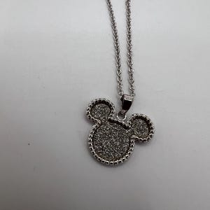K&ouml;nnte beinhalten: Silberfarbene Halskette mit einem Anh&auml;nger in Form eines M&auml;usekopfes. Der Anh&auml;nger hat eine strukturierte, glitzernde Oberfl&auml;che und ist mit kleinen, perlenartigen Details umrandet. Die Kette ist filigran und h&auml;ngt an einem kleinen Verschluss.