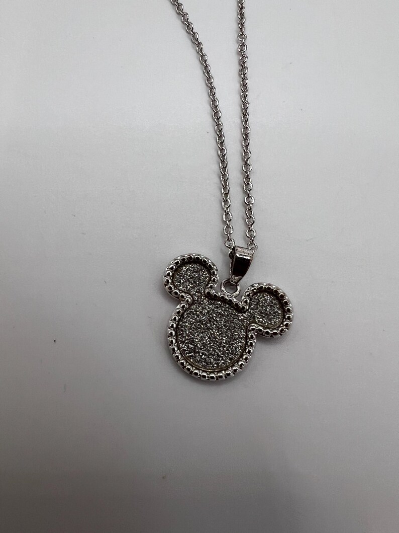 K&ouml;nnte beinhalten: Silberfarbene Halskette mit einem Anh&auml;nger in Form eines M&auml;usekopfes. Der Anh&auml;nger hat eine strukturierte, glitzernde Oberfl&auml;che und ist mit kleinen, perlenartigen Details umrandet. Die Kette ist filigran und h&auml;ngt an einem kleinen Verschluss.