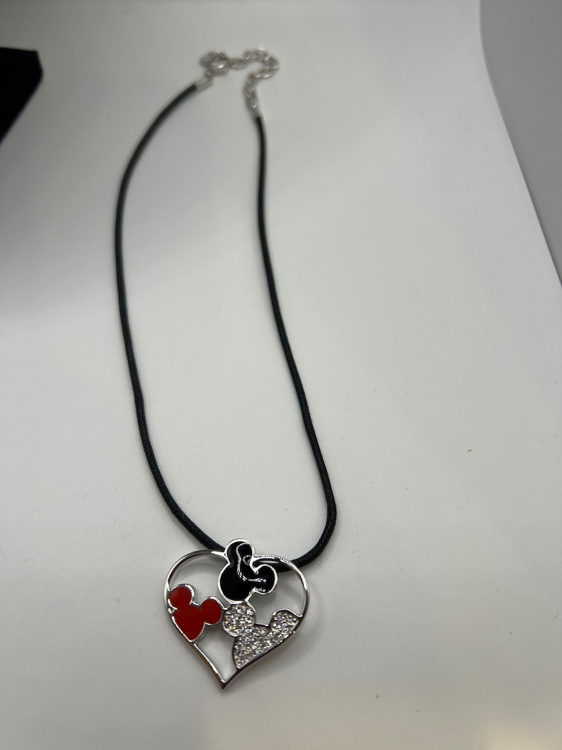 Puede incluir: Un collar de cord&oacute;n negro con un colgante de coraz&oacute;n plateado con un dise&ntilde;o de Mickey Mouse rojo y negro con pedrer&iacute;a.