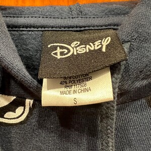 Pu&ograve; includere: Una felpa con cappuccio blu navy con una grafica bianca della testa di Topolino. L'etichetta dice "Disney" e elenca il contenuto del tessuto come 60% cotone e 40% poliestere. L'etichetta dice anche "RN# 117508" e "Made in China". L'etichetta della taglia dice "S".