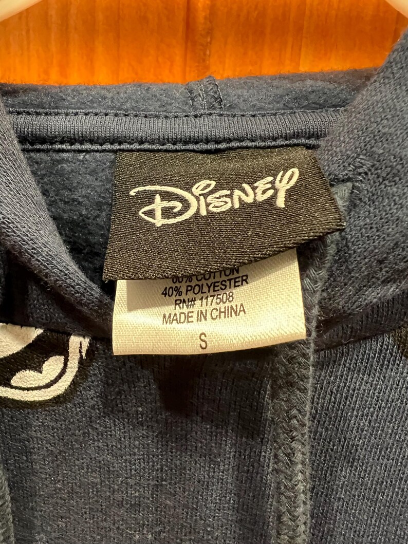 Pu&ograve; includere: Una felpa con cappuccio blu navy con una grafica bianca della testa di Topolino. L'etichetta dice "Disney" e elenca il contenuto del tessuto come 60% cotone e 40% poliestere. L'etichetta dice anche "RN# 117508" e "Made in China". L'etichetta della taglia dice "S".