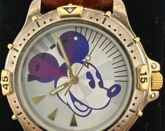 May include: A silver and gold wristwatch with a brown leather band. The watch face features a white background with a Mickey Mouse design and black numbers around the edge.