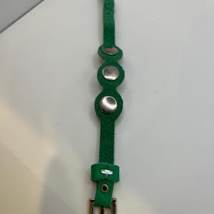 Puede incluir: Una pulsera de cuero verde con tres remaches plateados y una hebilla plateada. La pulsera est&aacute; hecha de una sola tira de cuero verde con una serie de agujeros para ajustar. La hebilla es rectangular y de metal plateado.