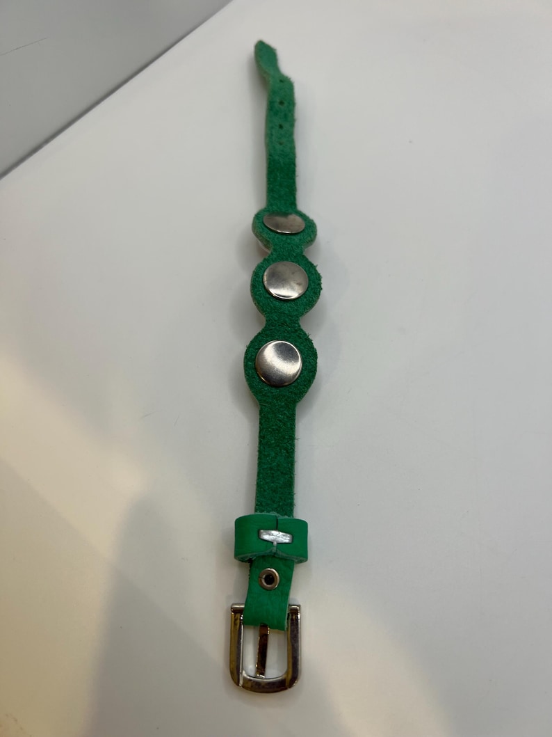 Puede incluir: Una pulsera de cuero verde con tres remaches plateados y una hebilla plateada. La pulsera est&aacute; hecha de una sola tira de cuero verde con una serie de agujeros para ajustar. La hebilla es rectangular y de metal plateado.