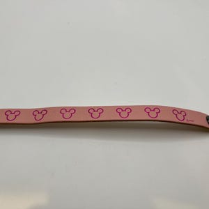 Puede incluir: Una pulsera de cuero rosa claro con un patr&oacute;n repetido de siluetas de la cabeza de Mickey Mouse en rosa m&aacute;s oscuro. La pulsera tiene un cierre de bot&oacute;n a presi&oacute;n de metal y un remache decorativo de metal.