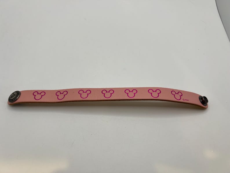 Puede incluir: Una pulsera de cuero rosa claro con un patr&oacute;n repetido de siluetas de la cabeza de Mickey Mouse en rosa m&aacute;s oscuro. La pulsera tiene un cierre de bot&oacute;n a presi&oacute;n de metal y un remache decorativo de metal.