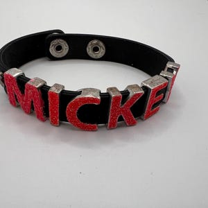 Puede incluir: Pulsera de cuero negro con el nombre "MICKEY" escrito en letras rojas con purpurina y bordes de metal plateado. La pulsera tiene un cierre de bot&oacute;n a presi&oacute;n.