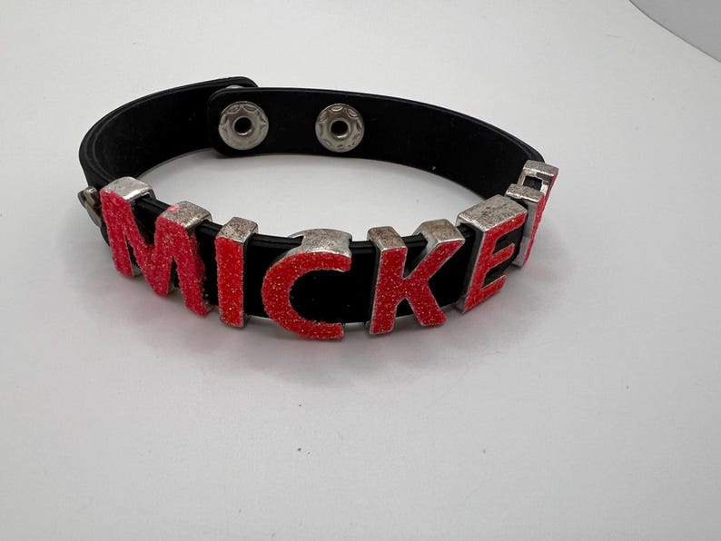 Puede incluir: Pulsera de cuero negro con el nombre "MICKEY" escrito en letras rojas con purpurina y bordes de metal plateado. La pulsera tiene un cierre de bot&oacute;n a presi&oacute;n.