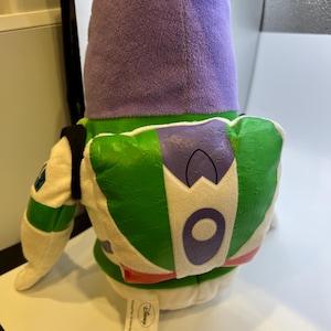 May include: A plush toy of Buzz Lightyear from the Toy Story franchise. The toy is wearing a purple hooded jacket and has a green and white spacesuit with a purple chest emblem. The toy is sitting on a white surface with a Disney label.