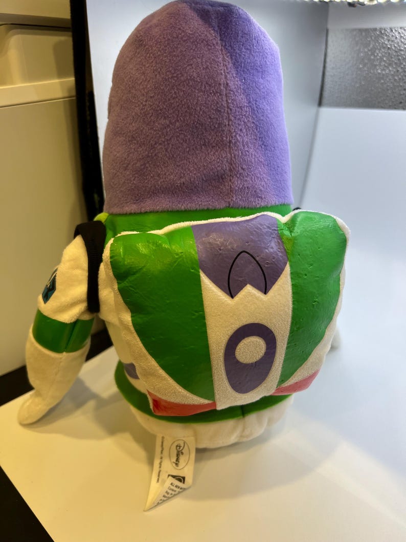 May include: A plush toy of Buzz Lightyear from the Toy Story franchise. The toy is wearing a purple hooded jacket and has a green and white spacesuit with a purple chest emblem. The toy is sitting on a white surface with a Disney label.