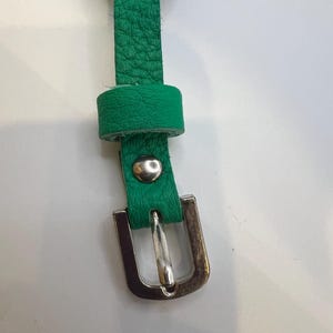 Puede incluir: Primer plano de una correa de cuero verde con una hebilla de metal plateado. La correa tiene una superficie texturizada y un peque&ntilde;o lazo. Un remache plateado asegura la correa a la hebilla.