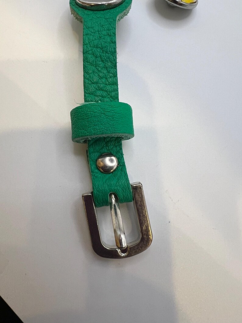 Puede incluir: Primer plano de una correa de cuero verde con una hebilla de metal plateado. La correa tiene una superficie texturizada y un peque&ntilde;o lazo. Un remache plateado asegura la correa a la hebilla.