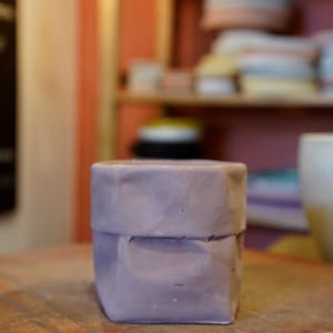 Puede incluir: Un recipiente de cerámica cuadrado de color lavanda con una parte superior doblada, que se asemeja a una bolsa de papel. El recipiente está sobre una superficie redonda de madera con un patrón de vetas de madera natural. El fondo está borroso, mostrando estantes y otros objetos.