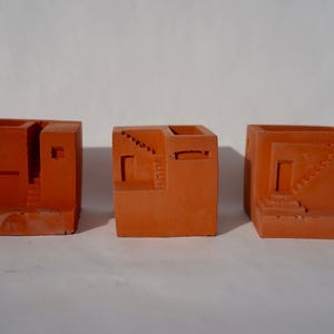 May include: Three orange-colored, cube-shaped planters with architectural designs. Each planter features cut-out details resembling miniature buildings with stairs, doors, and windows. The planters are arranged on a white surface.