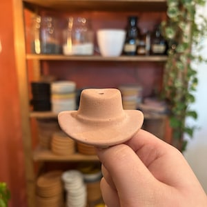 Puede incluir: Un pequeño sombrero de vaquero de color terracota. El sombrero se sostiene en una mano y tiene un ala ancha y una corona redondeada. El sombrero es probablemente un artículo decorativo o un pequeño accesorio.