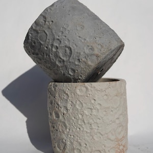 May include: Two concrete planters with a textured, cratered surface resembling the moon. One is light gray, the other is dark gray, and they are stacked. The planters are cylindrical and suitable for indoor or outdoor use.