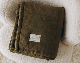 May include: A dark green quilted throw blanket with a white label that reads "Adamo Joya 100% French Flax Linen" on a white fluffy surface.