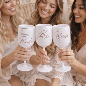 Puede incluir: Tres copas de vino blancas con letras doradas que dicen "Team Bride", "Alex" y "Bride". Las copas son sostenidas por tres personas. La imagen está tomada en una habitación bien iluminada, con un fondo suave y neutro. El tema es nupcial.