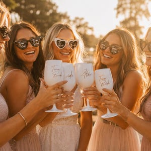 Puede incluir: Un grupo de mujeres con gafas de sol en forma de corazón y sosteniendo copas de vino blanco. Las copas están inscritas con "Team Bride" y un nombre. Las mujeres visten vestidos de colores claros, celebrando al aire libre bajo el sol.