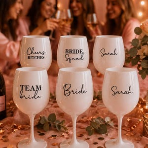 Puede incluir: Seis copas de vino blancas con texto negro, incluyendo "Cheers Bitches", "Bride Squad", "Sarah", "Team Bride" y "Bride". También se ve una botella de champán Moët & Chandon y una vela encendida. La escena está ambientada sobre una superficie rosa con decoraciones florales.