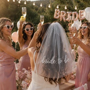 Könnte beinhalten: Ein weißer Brautschleier mit verstreuten kleinen Perlen und dem in weißer Schreibschrift gestickten Text bride to be. Der Schleier besteht aus transparentem Tüllstoff und wird von einer Person mit langem dunklem Haar getragen.