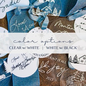 May include: A selection of clear and white round acrylic baubles featuring various Christmas-themed designs and text, such as "Merry Christmas", "Love You", "Count It All Joy", and "Holy Night". The baubles are arranged on a wooden surface with blue ribbon.