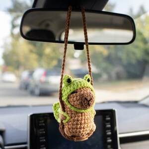 Puede incluir: Un adorno de coche de ganchillo hecho a mano con un cuerpo marrón y un gorro de rana verde. El adorno está suspendido de un cordón marrón y cuelga del espejo retrovisor de un coche. El gorro de rana tiene ojos negros y una cara sonriente.
