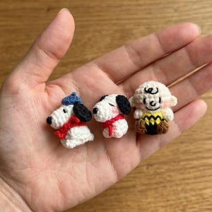 May include: Three small, handmade crochet figures of Snoopy and Charlie Brown characters. Snoopy is wearing a blue hat and red scarf. Charlie Brown has a yellow shirt with a zig-zag pattern and black shorts. The figures are white, black, red, blue, and brown.