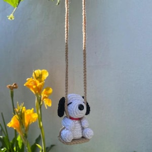 K&ouml;nnte beinhalten: Ein handgeh&auml;kelter Snoopy-Hund auf einer Schaukel. Der wei&szlig;e Snoopy hat schwarze Ohren und ein rotes Halsband. Die Schaukel ist aus beigem Garn gefertigt und an einem beigen Seil aufgeh&auml;ngt. Gelbe Blumen und gr&uuml;ne Bl&auml;tter sind im Hintergrund.