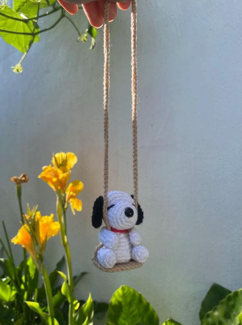 K&ouml;nnte beinhalten: Ein handgeh&auml;kelter Snoopy-Hund auf einer Schaukel. Der wei&szlig;e Snoopy hat schwarze Ohren und ein rotes Halsband. Die Schaukel ist aus beigem Garn gefertigt und an einem beigen Seil aufgeh&auml;ngt. Gelbe Blumen und gr&uuml;ne Bl&auml;tter sind im Hintergrund.