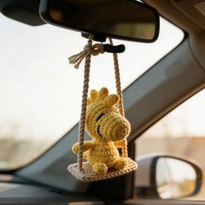 K&ouml;nnte beinhalten: Ein gelber, geh&auml;kelter Woody-Vogel-Autoanh&auml;nger, der am R&uuml;ckspiegel eines Autos h&auml;ngt. Der Anh&auml;nger ist an einer beigefarbenen Seilschaukel befestigt. Der Innenraum des Autos ist sichtbar.