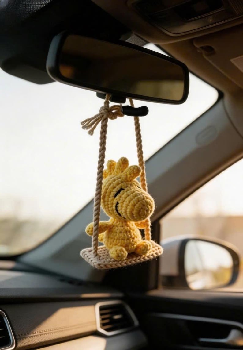 K&ouml;nnte beinhalten: Ein gelber, geh&auml;kelter Woody-Vogel-Autoanh&auml;nger, der am R&uuml;ckspiegel eines Autos h&auml;ngt. Der Anh&auml;nger ist an einer beigefarbenen Seilschaukel befestigt. Der Innenraum des Autos ist sichtbar.