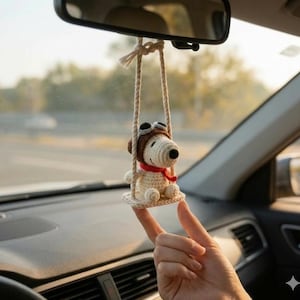 K&ouml;nnte beinhalten: Geh&auml;kelter Snoopy-Autoanh&auml;nger, der an einem R&uuml;ckspiegel h&auml;ngt. Der Anh&auml;nger zeigt Snoopy mit braunem Fliegerm&uuml;tze und rotem Schal, der auf einer Schaukel sitzt. Die Schaukel besteht aus beigefarbenem Garn. Der Hintergrund ist verschwommen und zeigt einen Autoinnenraum und eine Au&szlig;enansicht.