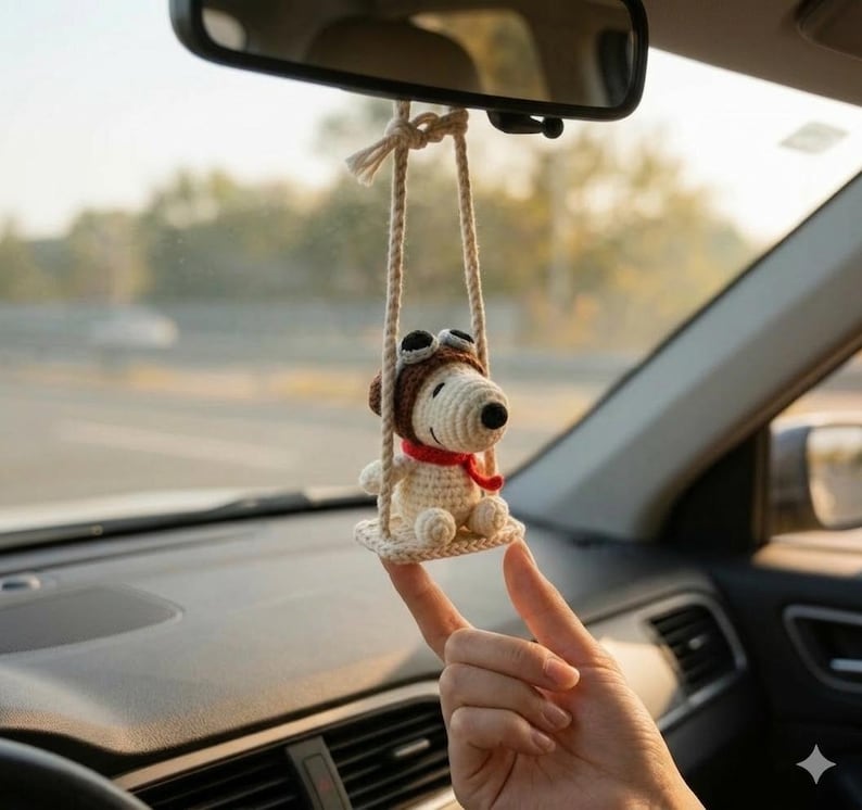 K&ouml;nnte beinhalten: Geh&auml;kelter Snoopy-Autoanh&auml;nger, der an einem R&uuml;ckspiegel h&auml;ngt. Der Anh&auml;nger zeigt Snoopy mit braunem Fliegerm&uuml;tze und rotem Schal, der auf einer Schaukel sitzt. Die Schaukel besteht aus beigefarbenem Garn. Der Hintergrund ist verschwommen und zeigt einen Autoinnenraum und eine Au&szlig;enansicht.