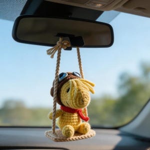 K&ouml;nnte beinhalten: Ein gelber geh&auml;kelter Charakter mit brauner Fliegerm&uuml;tze und rotem Schal, der am R&uuml;ckspiegel eines Autos h&auml;ngt. Die Figur sitzt auf einer kleinen, beigen Schaukel und verleiht dem Innenraum des Autos einen Hauch von Verspieltheit.