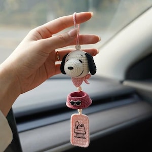 May include: Crocheted Snoopy car charm. The charm features Snoopy's head with black ears and a pink bow, a pink bowl, and a pink tag with a Snoopy graphic. The charm is suspended by a pink string and is held in a hand.