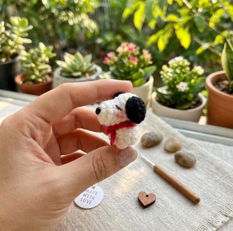 K&ouml;nnte beinhalten: Eine kleine, handgefertigte H&auml;kelhundfigur in Wei&szlig;, Schwarz und Rot. Der Hund hat schwarze Ohren und einen schwarzen Fleck &uuml;ber einem Auge, mit einem roten Schal. Ein kleines Etikett lautet "MADE WITH LOVE".