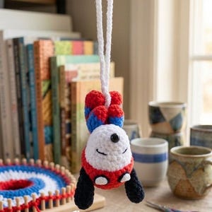 Op de afbeelding: Een gehaakte panda-ornament met een witte kop, rode en zwarte details en blauwe en rode accenten, hangend aan een wit touwtje. De achtergrond bevat boeken, mokken en een knutselproject. Een charmant handgemaakt item.